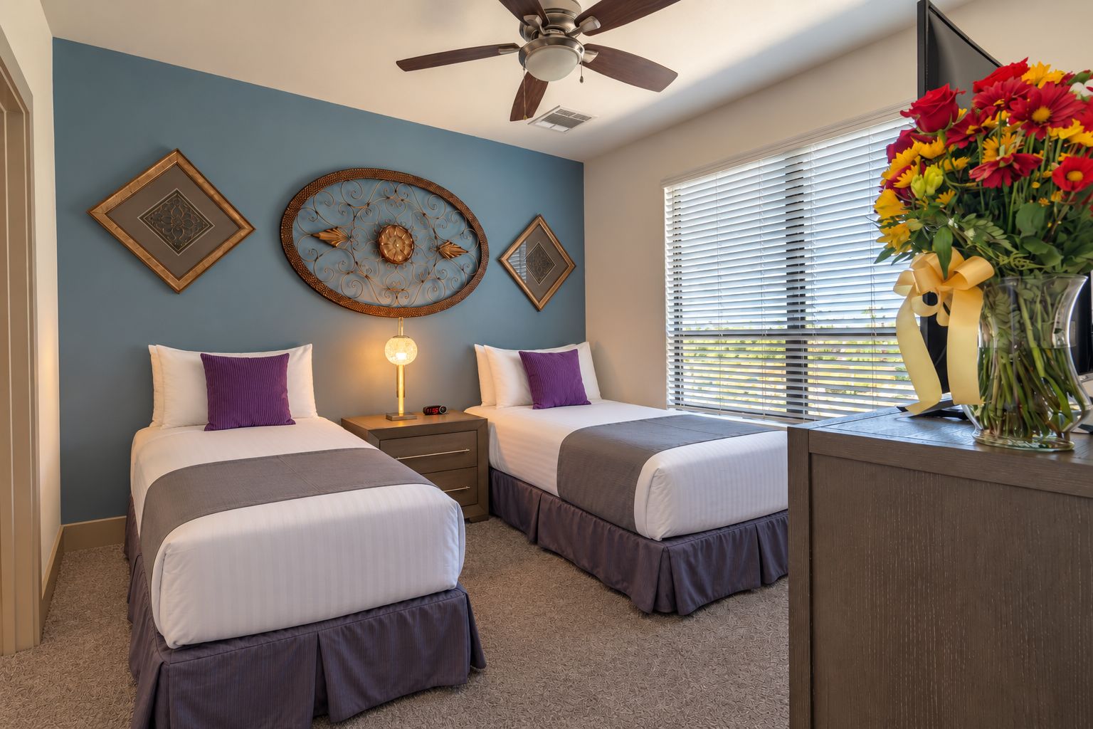 A hotel room with two beds, a ceiling fan, decorative wall art, a bedside lamp, and a vase with flowers.