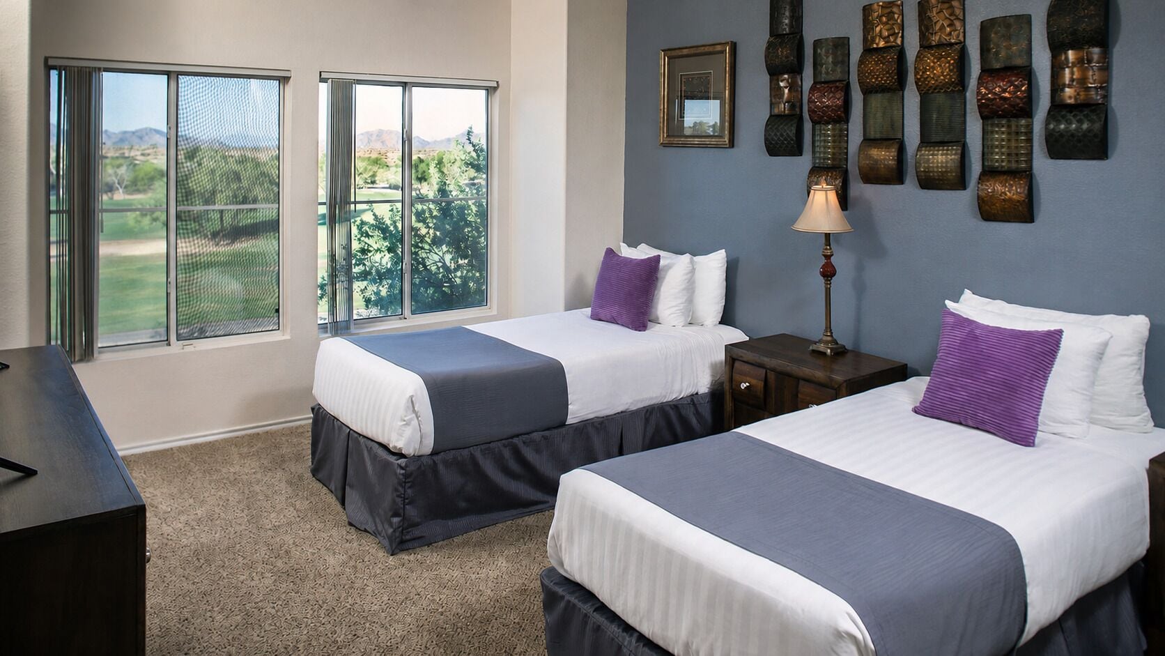 A bedroom with two beds, a desk, a lamp, and a wall with decorative items.