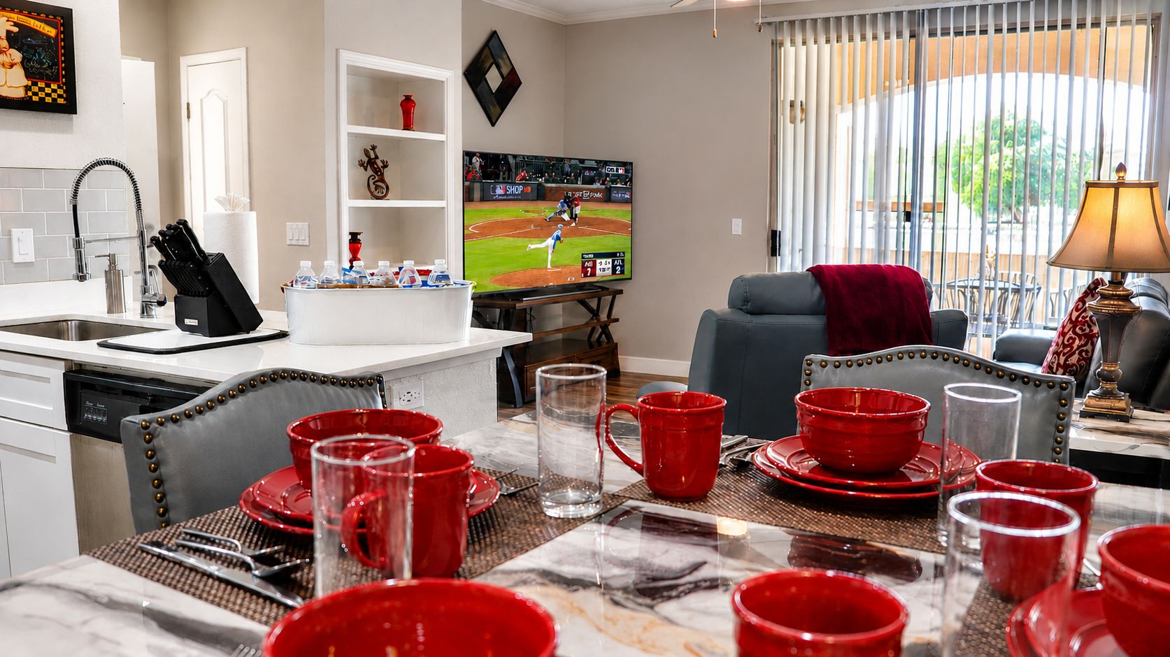 A dining area with a table set for four, a kitchen with a dishwasher, and a living room with a television displaying a baseball game.