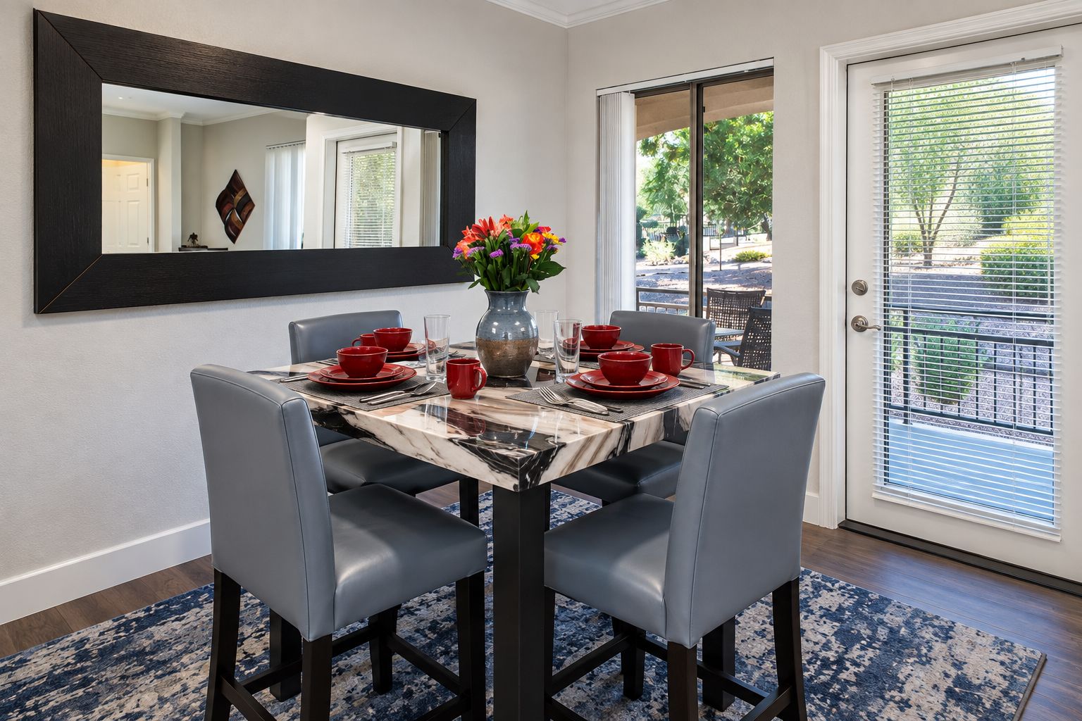 A dining area with a marble table, grey chairs, red tableware, and a vase of flowers.