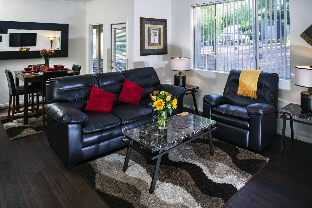 A modern living room with a black leather sofa, a glass coffee table, and a dining area with a wooden table and chairs.