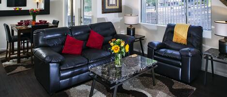 A modern living room with a black leather sofa, a glass coffee table, and a dining area with a wooden table and chairs.