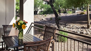 Un patio cubierto con una mesa y sillas, un jarrón con flores, y vista a un área ajardinada con árboles y rocas.