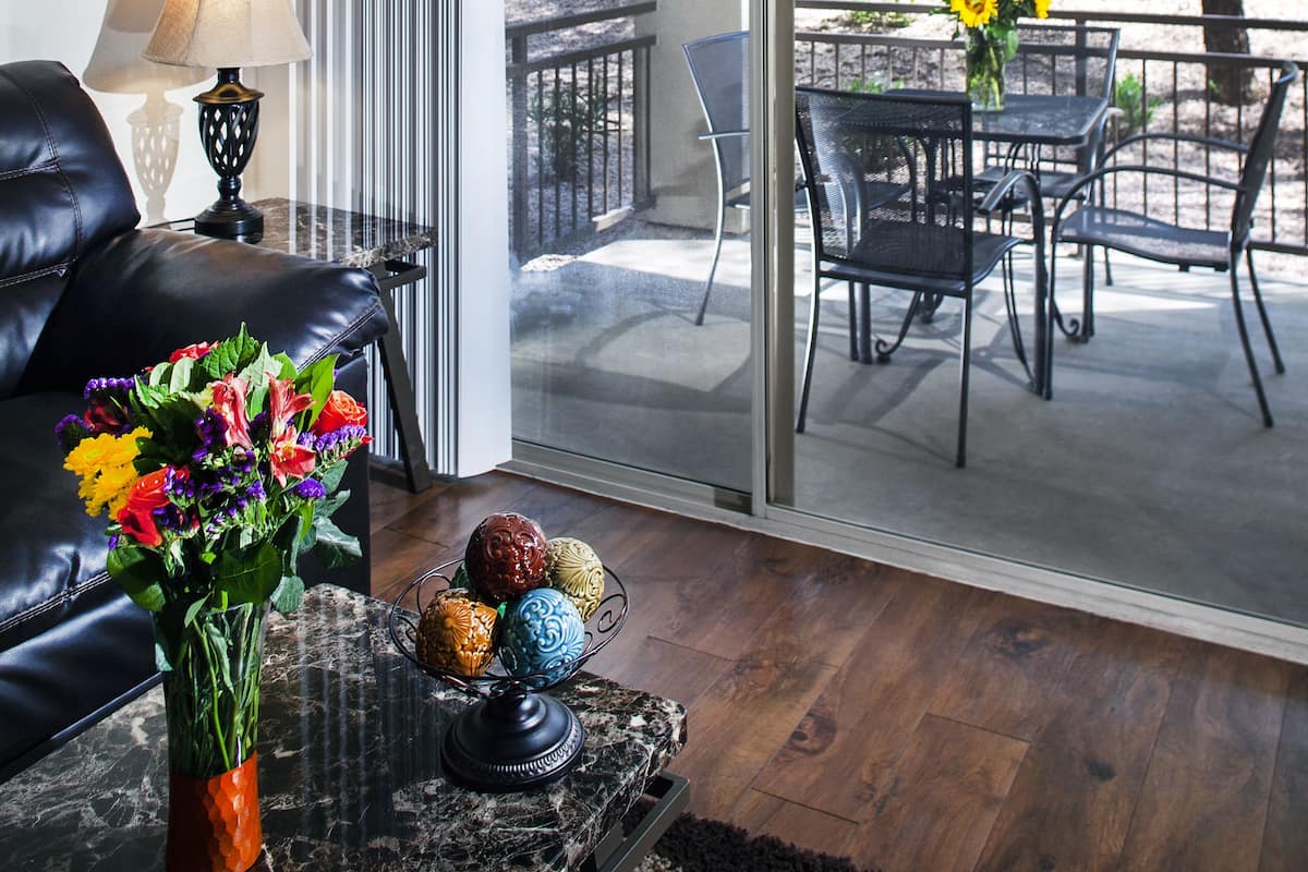 A modern living room with a leather sofa, a coffee table with a vase of flowers, and a glass door leading to an outdoor patio with a table and chairs.