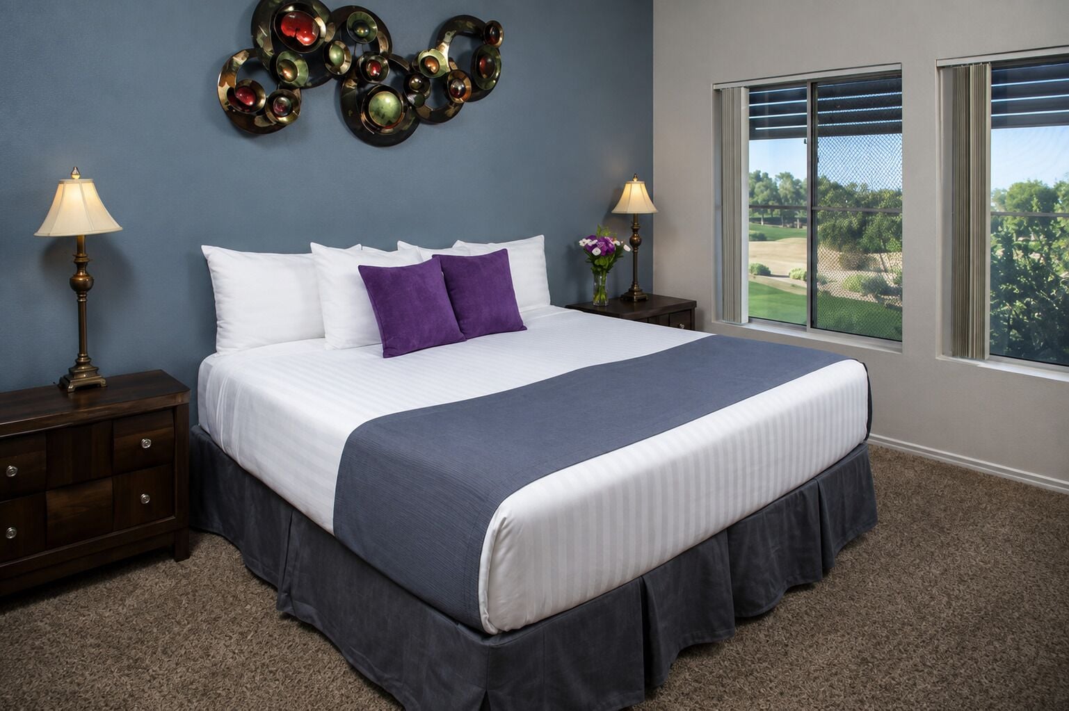 A bedroom with a large bed, two bedside tables with lamps, a decorative wall piece, and a window with a view of greenery.
