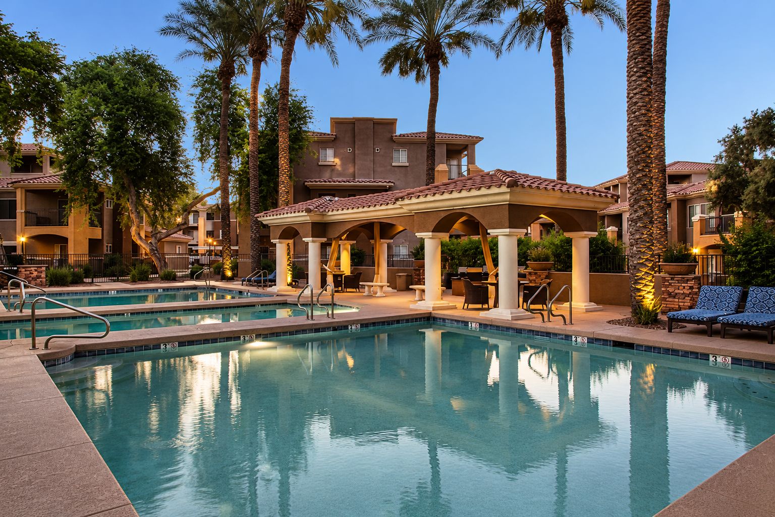 A pool area with a covered seating area, palm trees, and a building with balconies.