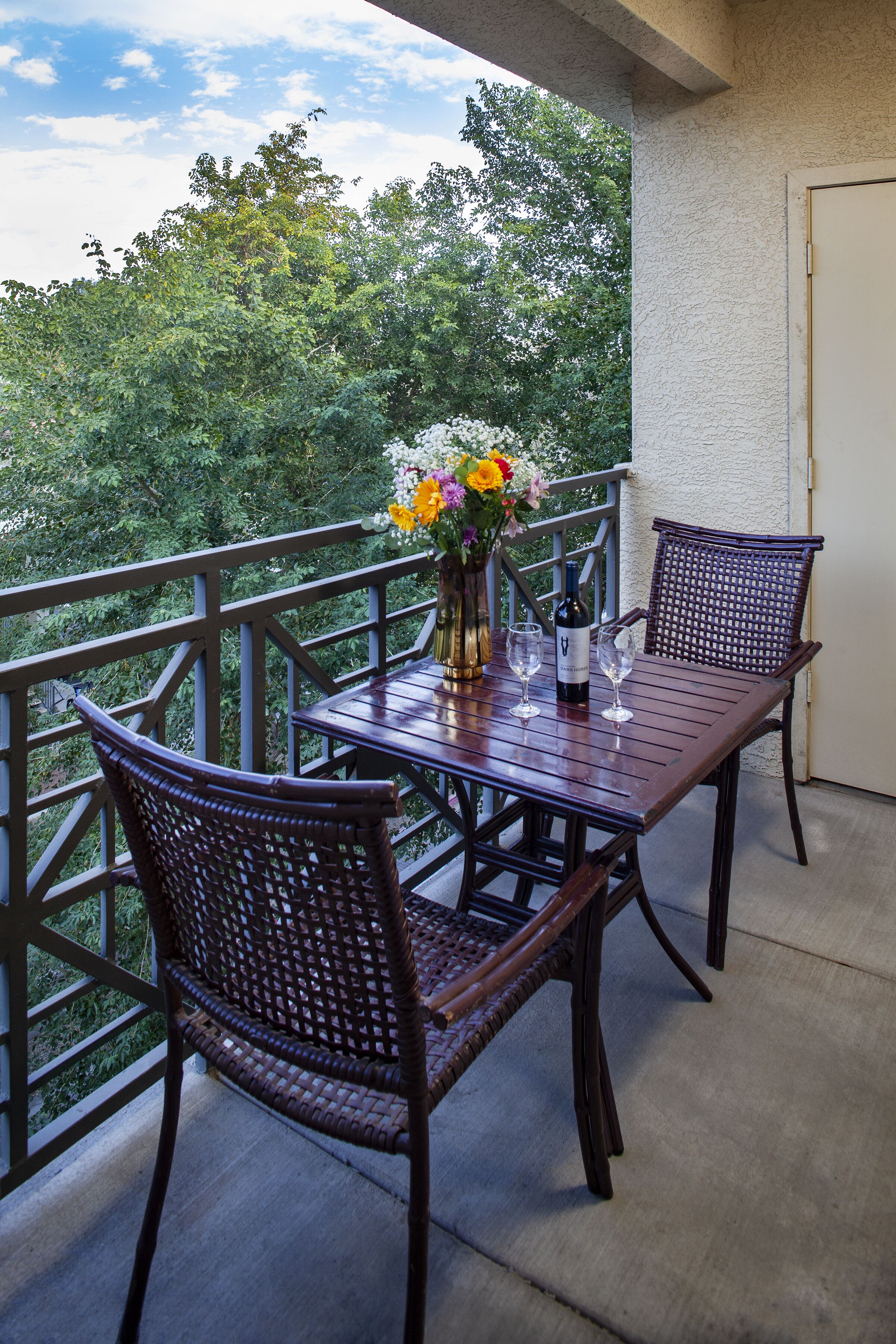 Ein Balkon mit einem Holztisch, Korbstühlen, einer Blumenvase und einer Weinflasche.