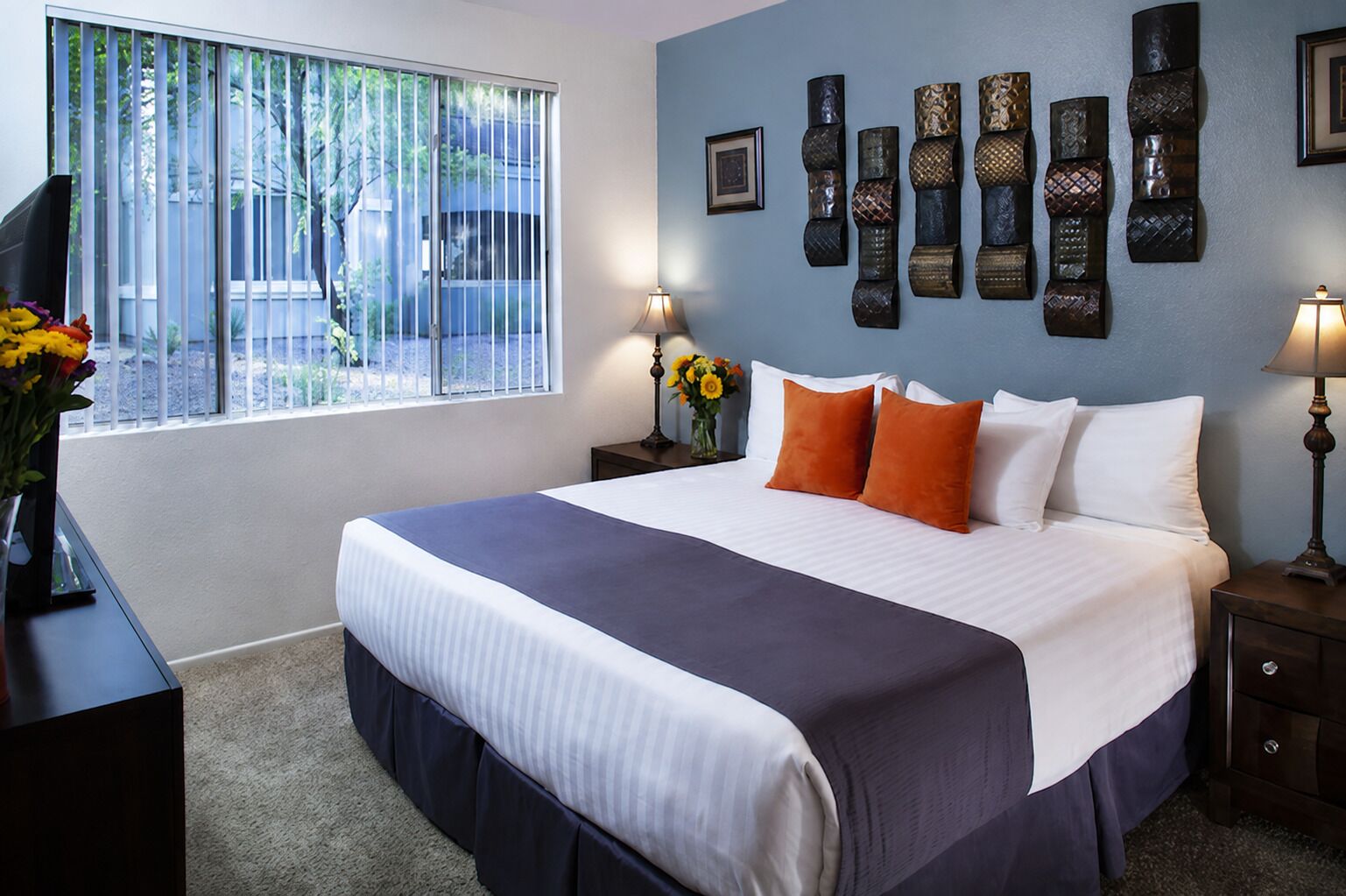 A bedroom with a large bed, two bedside tables, a window with blinds, and wall-mounted artwork.