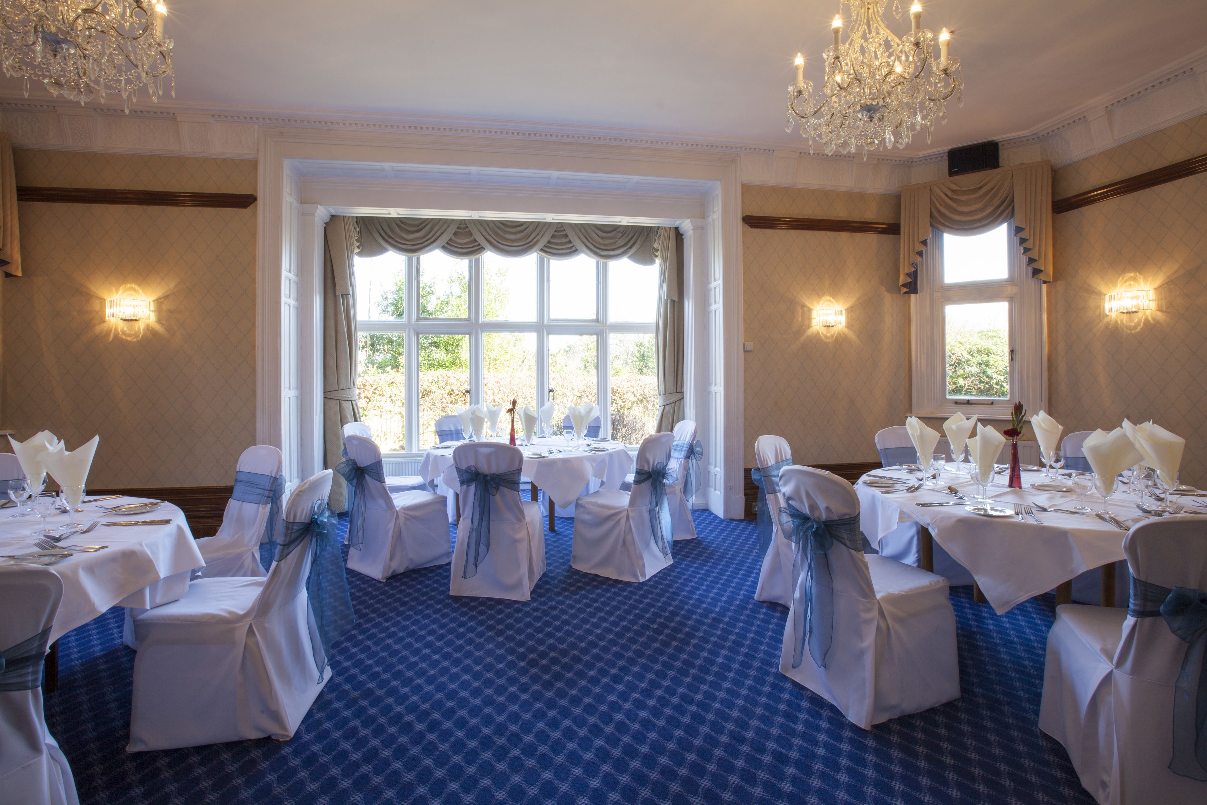 A banquet hall with tables set for a formal event, featuring chandeliers, large windows, and a patterned carpet.