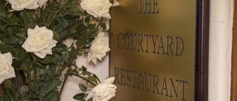 Letrero de 'The Courtyard Restaurant' con rosas blancas en un jarrón.