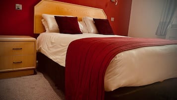 A neatly made bed with a red blanket and pillows, a wooden nightstand with drawers, and a wall-mounted light fixture.