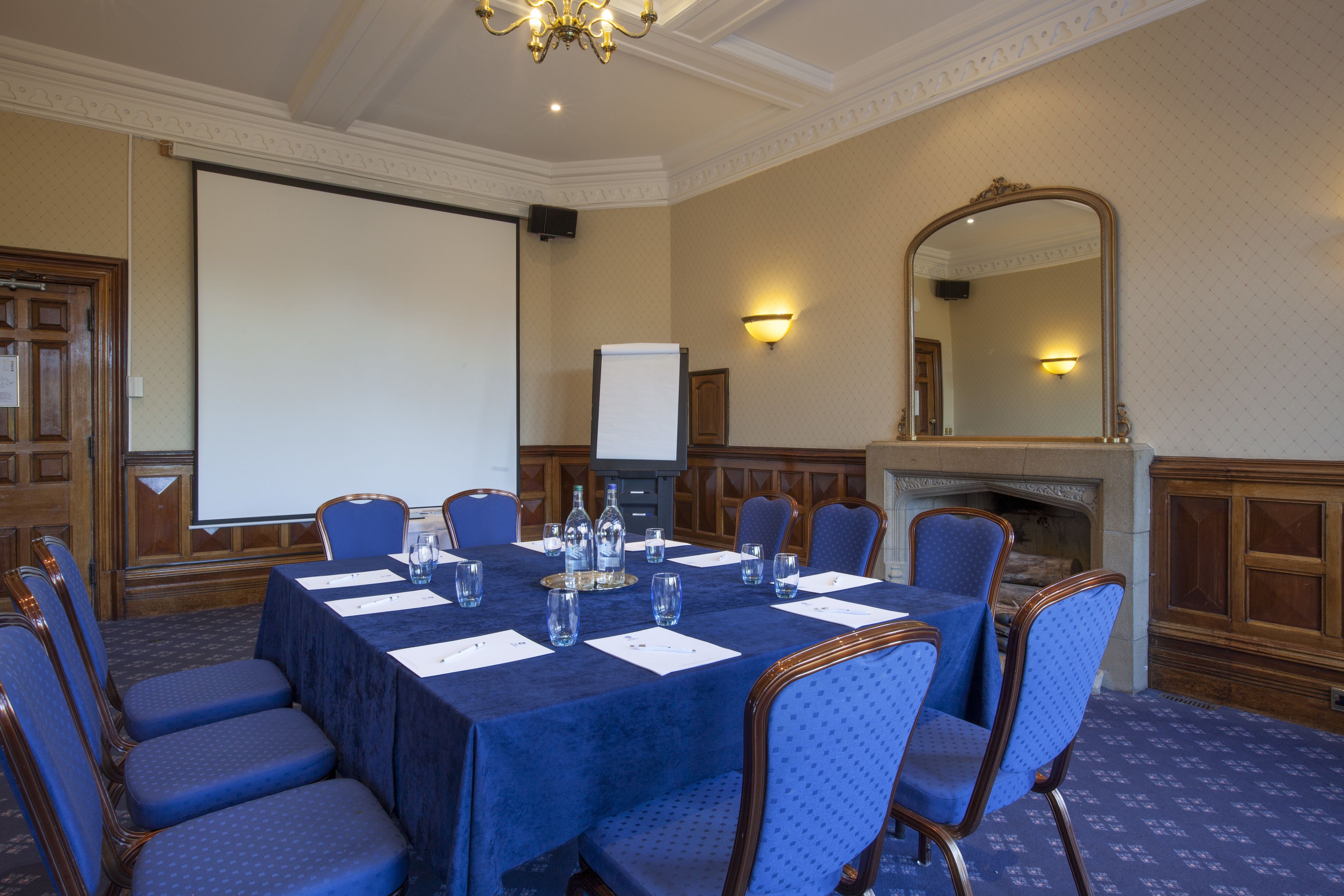 A conference room with a large table, blue chairs, a projector screen, a fireplace, and a mirror on the wall.