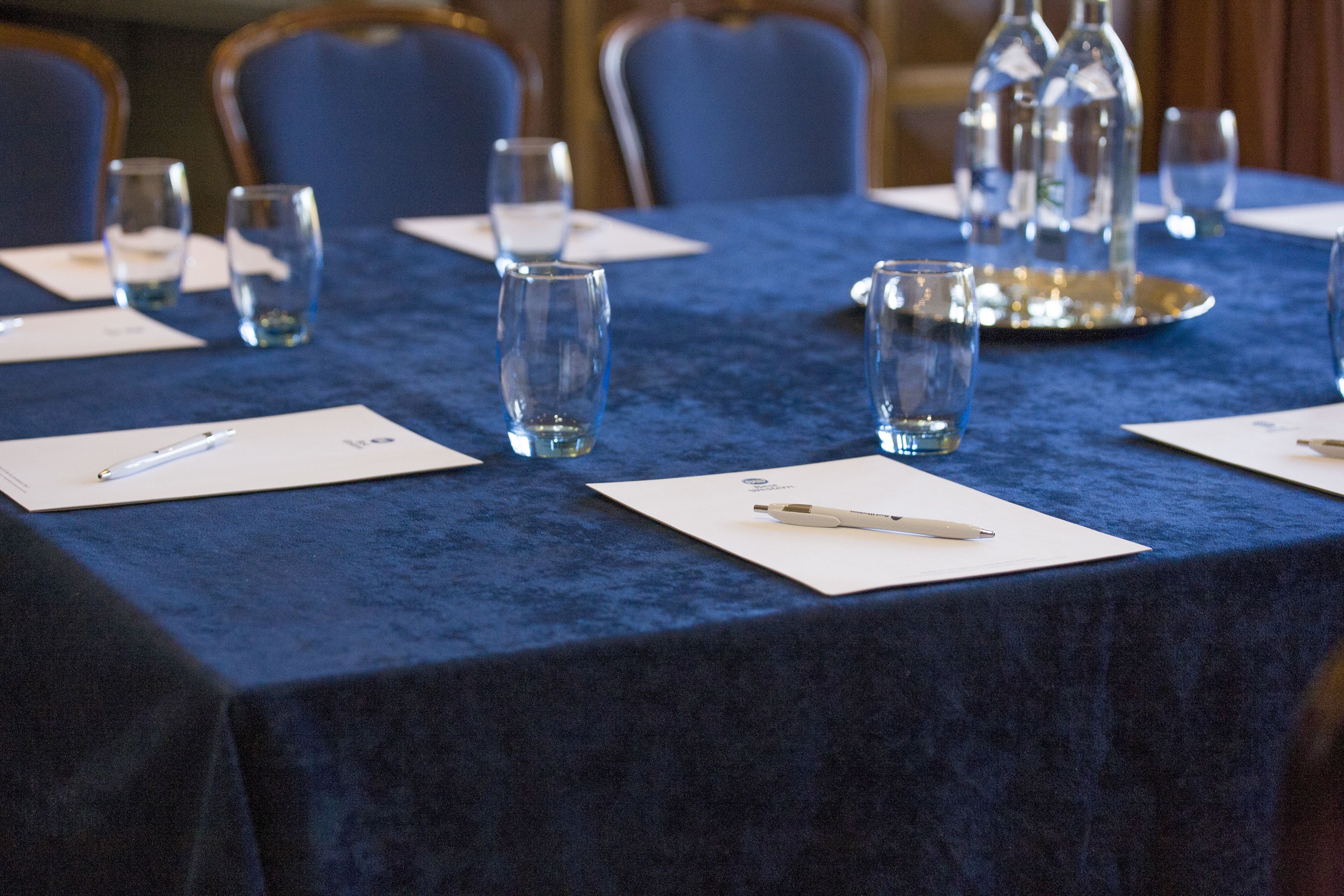 Une table de conférence, agrémentée de verres, de stylos et de cartes nominatives, laisse supposer qu’il s’agit d’une réunion ou d’un événement formel.