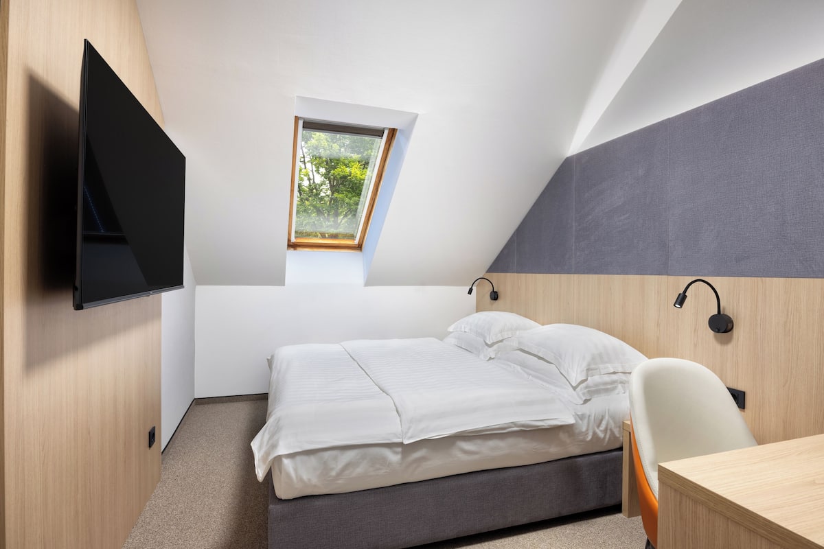Quarto moderno com cama, escrivaninha, cadeira, televisão na parede e janela com vista para árvores.