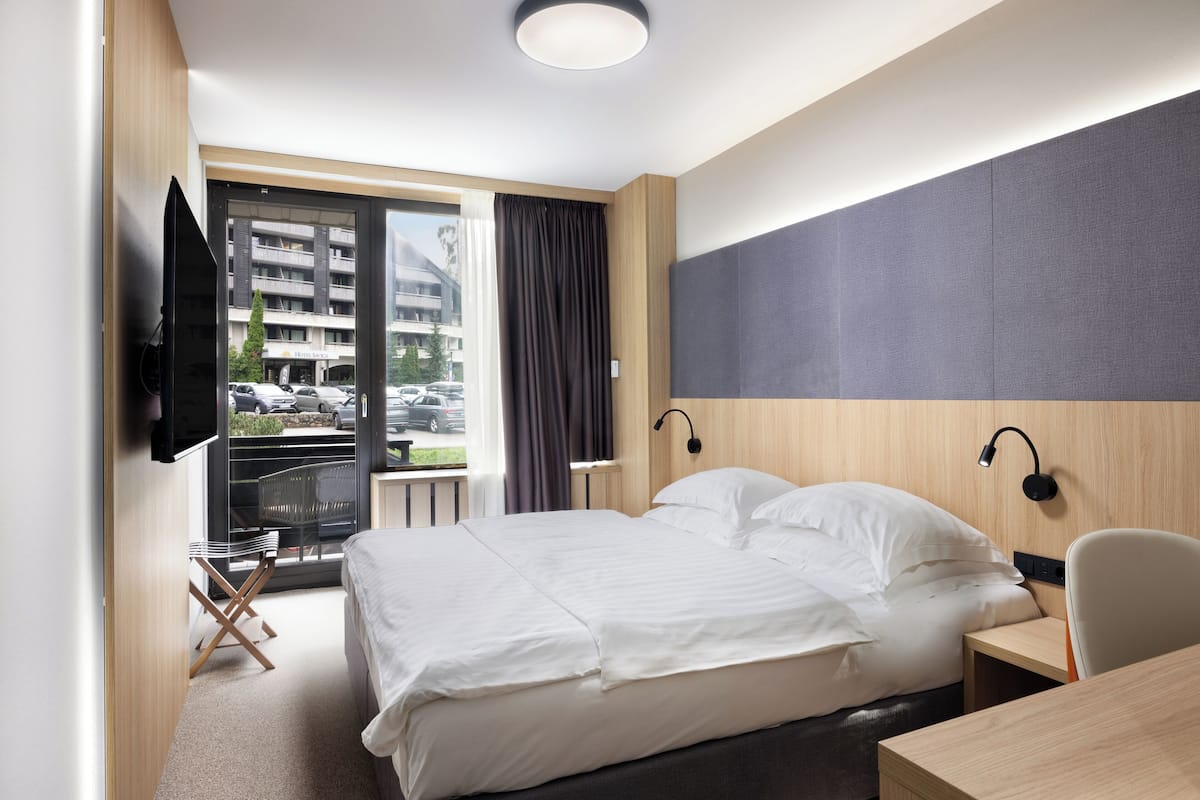 Quarto de hotel moderno com uma cama grande, uma mesa de trabalho, uma cadeira, televisão e varanda com vista para a rua.