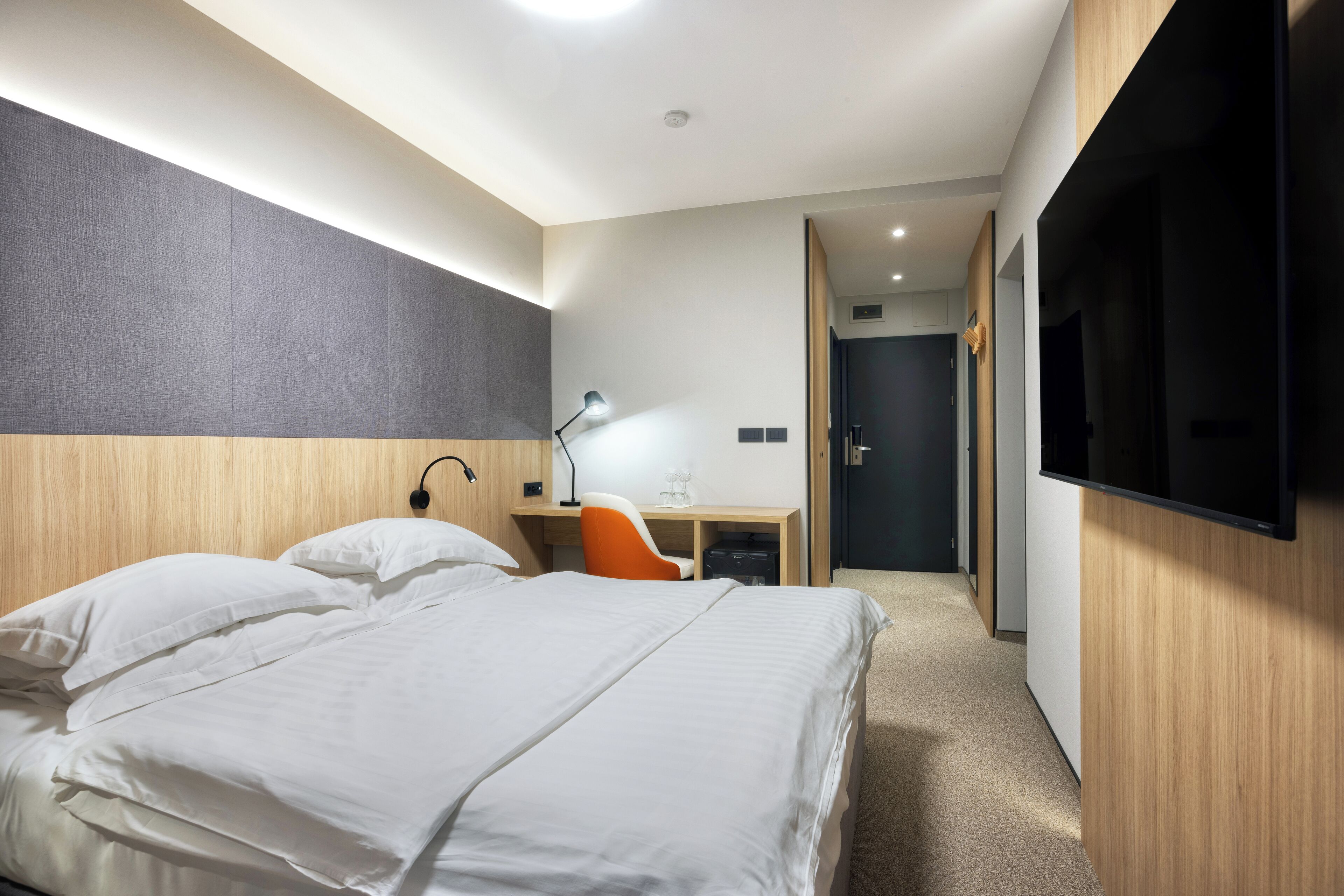 A hotel room with a bed, a desk, an orange chair, a desk lamp, a flat-screen TV, and a door leading to another room.