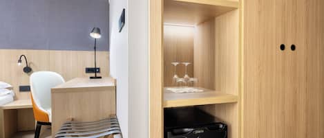 A compact hotel room with a wooden desk, a chair, a small kitchen area with a dishwasher, and a wardrobe with two doors.