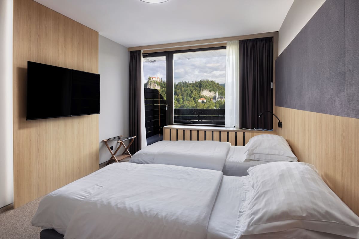 Quarto de hotel com duas camas, televisão presa na parede e uma grande janela com vista para um castelo e vegetação.