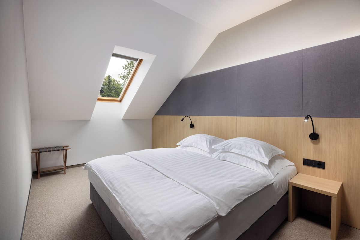Quarto com teto inclinado, claraboia, cama com roupa de cama branca, mesa de cabeceira e luminária fixada na parede.