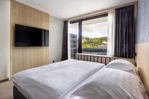 Relax in a serene space with a large bed, flat-screen TV, and a balcony view of lush greenery.