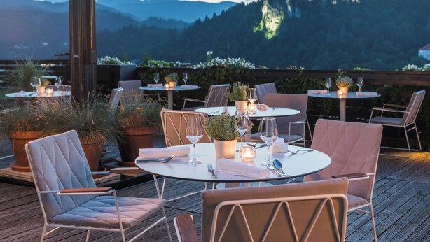 An outdoor dining area with tables and chairs, potted plants, and a view of mountains and a castle.