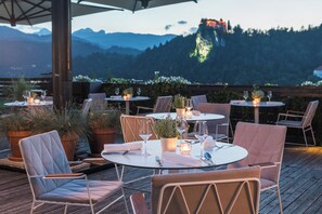 Un espace de restauration en plein air, avec des tables et des chaises, des plantes en pot et une vue sur les montagnes et un château.
