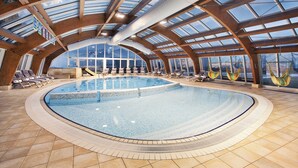 An indoor swimming pool with a curved design, surrounded by lounge chairs and hammocks, under a large glass roof.