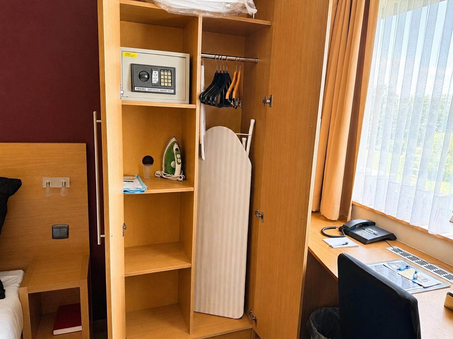 A compact hotel room with a bed, a desk with a computer, a telephone, and a window with blinds.