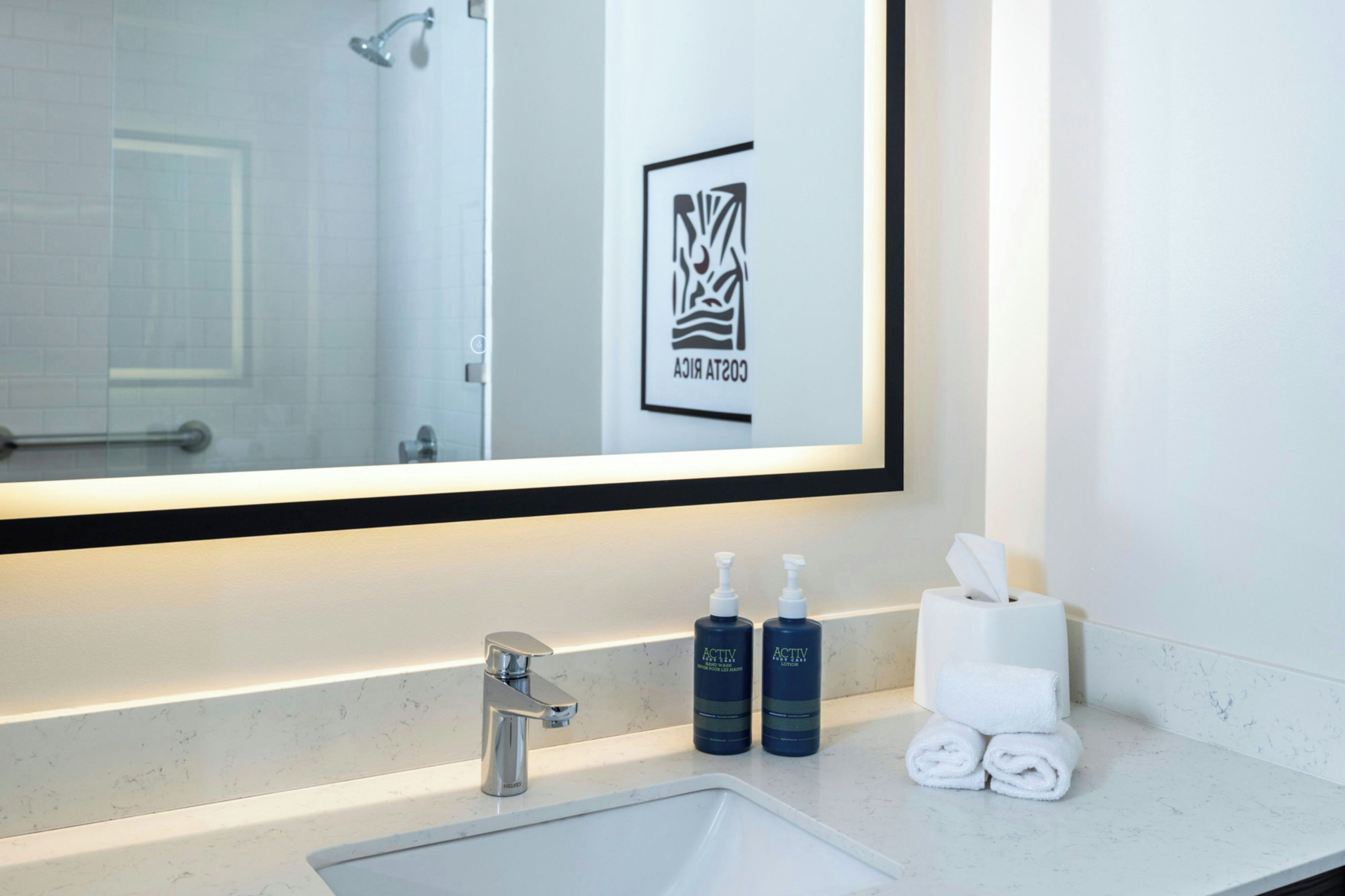 A hotel bathroom with a large mirror, a sink, two bottles of Activ body care lotion, a towel dispenser, and a framed logo on the wall.