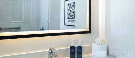 A hotel bathroom with a large mirror, a sink, two bottles of Activ body care lotion, a towel dispenser, and a framed logo on the wall.