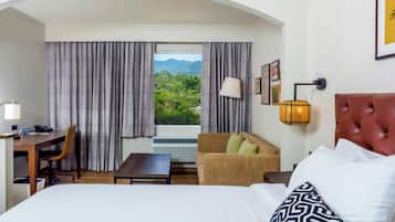 A hotel room with a bed, a desk, a chair, a sofa, and a view of greenery through the window.