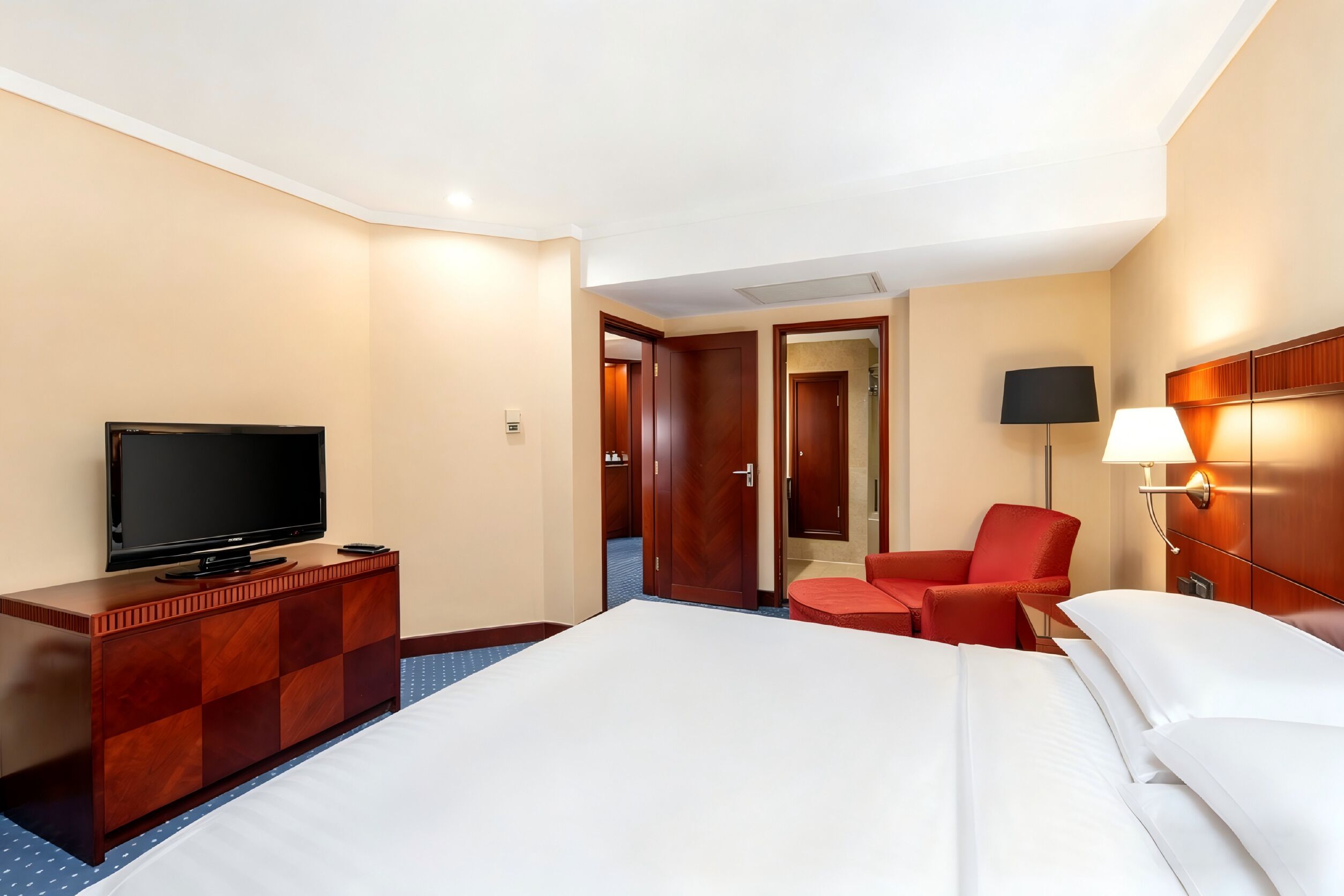 A hotel room with a bed, a TV on a wooden dresser, a red chair, a lamp, and a door leading to another room.