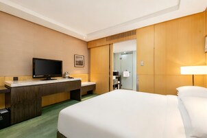 A hotel room with a bed, a desk with a flat-screen TV, a mirror, and a door leading to a bathroom.