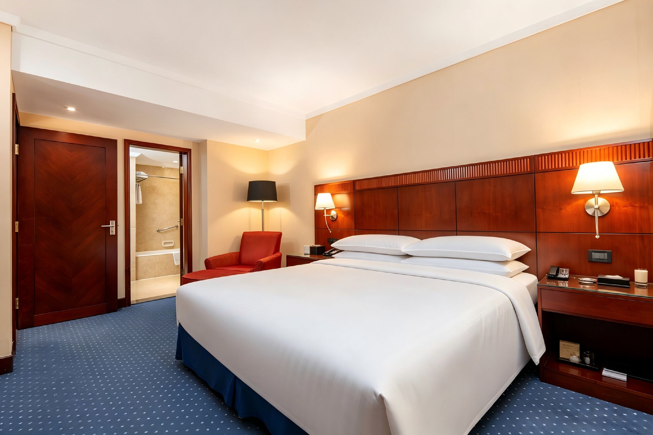 A hotel room with a large bed, a red chair, a wooden headboard, a nightstand, and a bathroom visible through an open door.