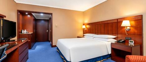 A hotel room with a bed, a desk with a television, a chair, a nightstand with a lamp, and a bathroom door.