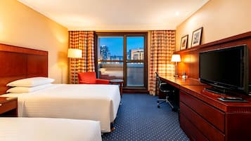 A hotel room with two beds, a desk with a TV, a chair, and a view of the cityscape through a large window.