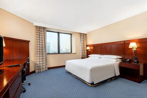 A hotel room with a bed, a desk, a chair, a TV, a nightstand with a lamp, and a window with curtains.