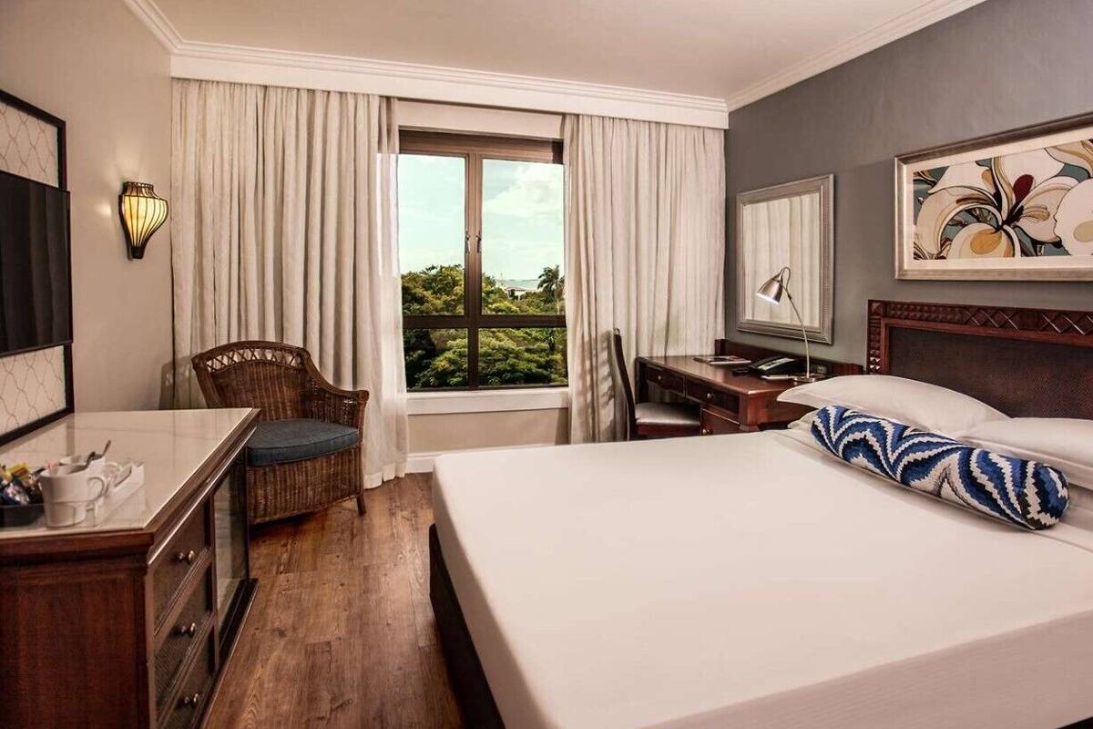 A hotel room with a bed, a desk, a chair, a window with curtains, a wall-mounted lamp, and a painting.