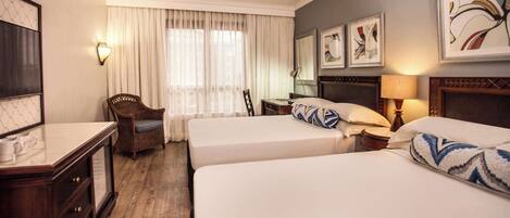A hotel room with two beds, a chair, a desk, a lamp, and artwork on the walls.