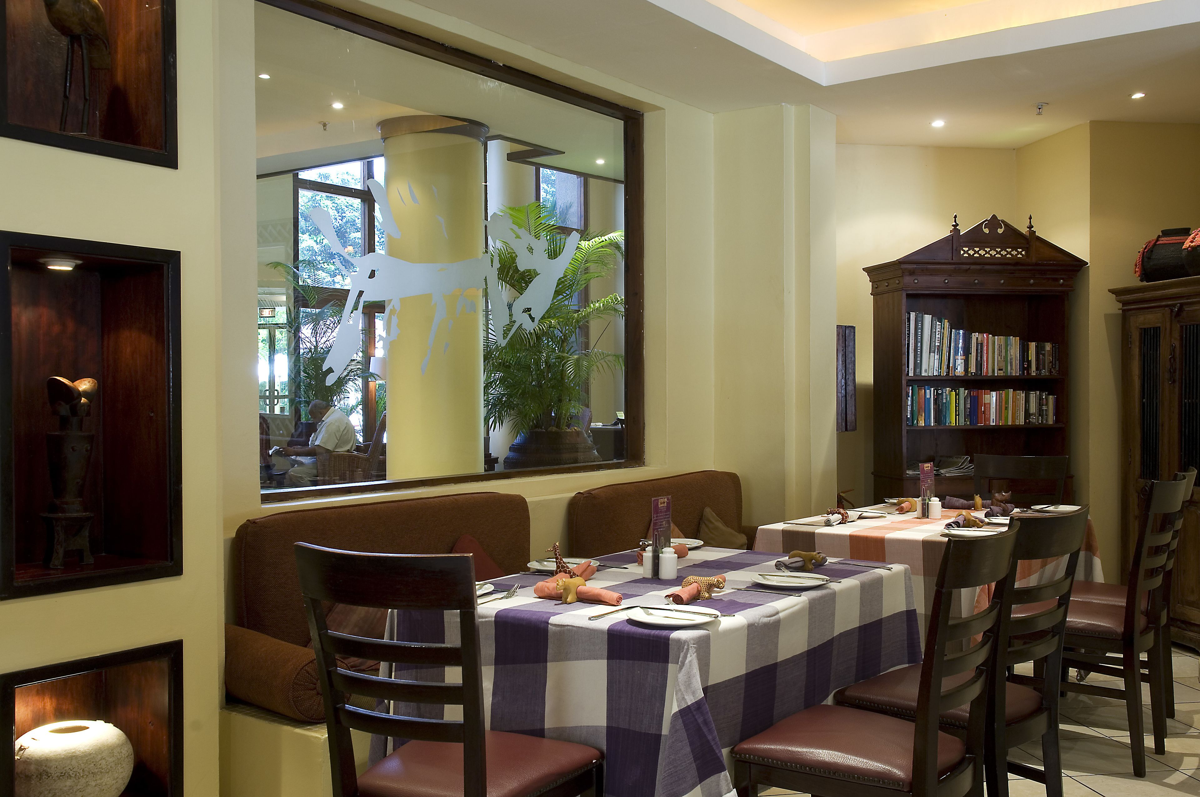 A dining area with tables set for two, a bookshelf, and a large window with a view of the outdoors.