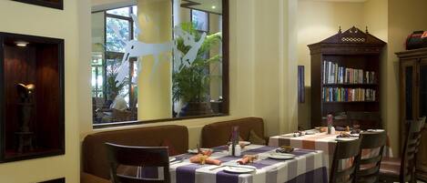 A dining area with tables set for two, a bookshelf, and a large window with a view of the outdoors.