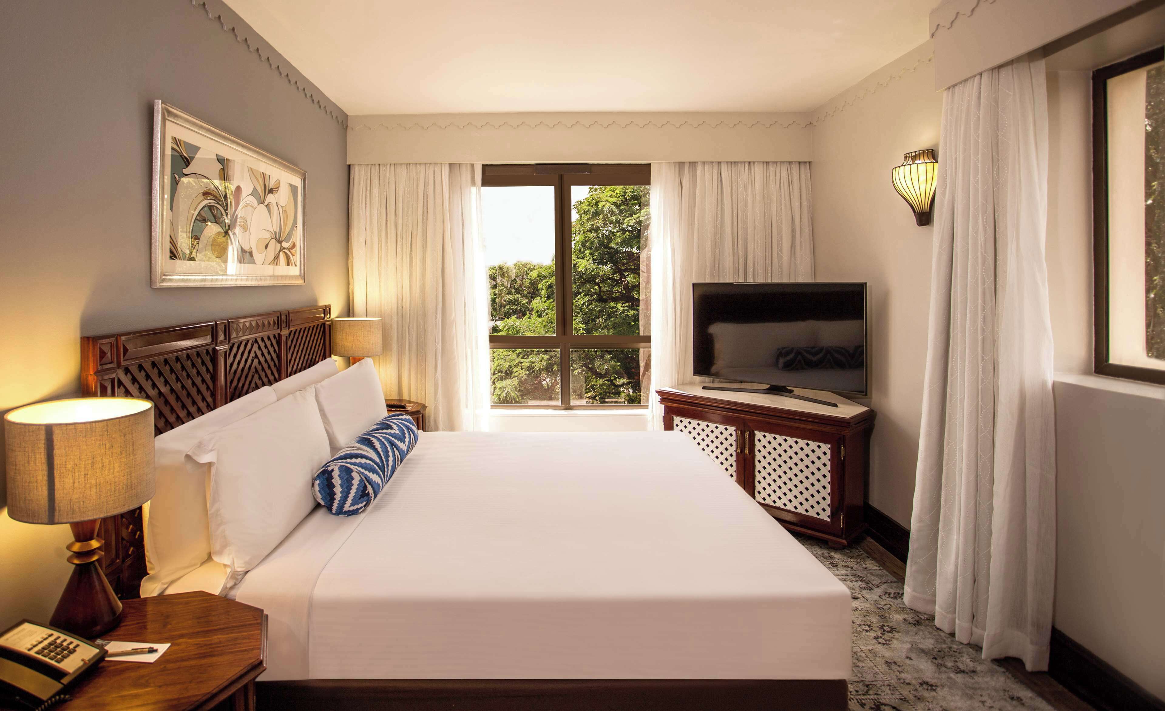 Una habitación de hotel con una cama grande, un televisor sobre un soporte de madera, un escritorio con una lámpara y una ventana con cortinas.