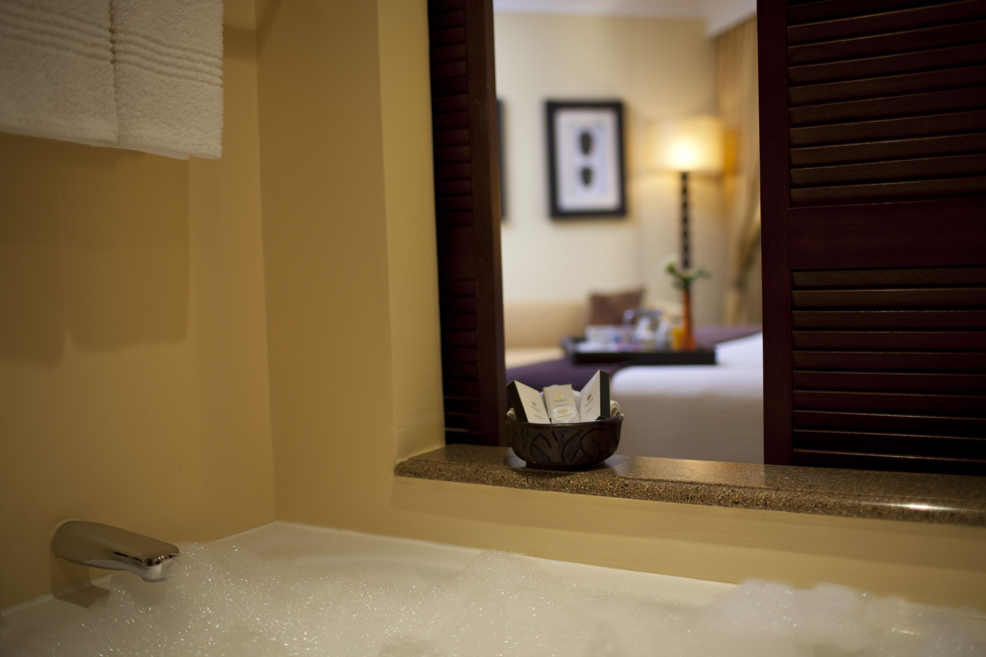 A bathroom with a bathtub filled with bubbles, a towel hanging on the wall, and a window with a view of a hotel room and a lamp.
