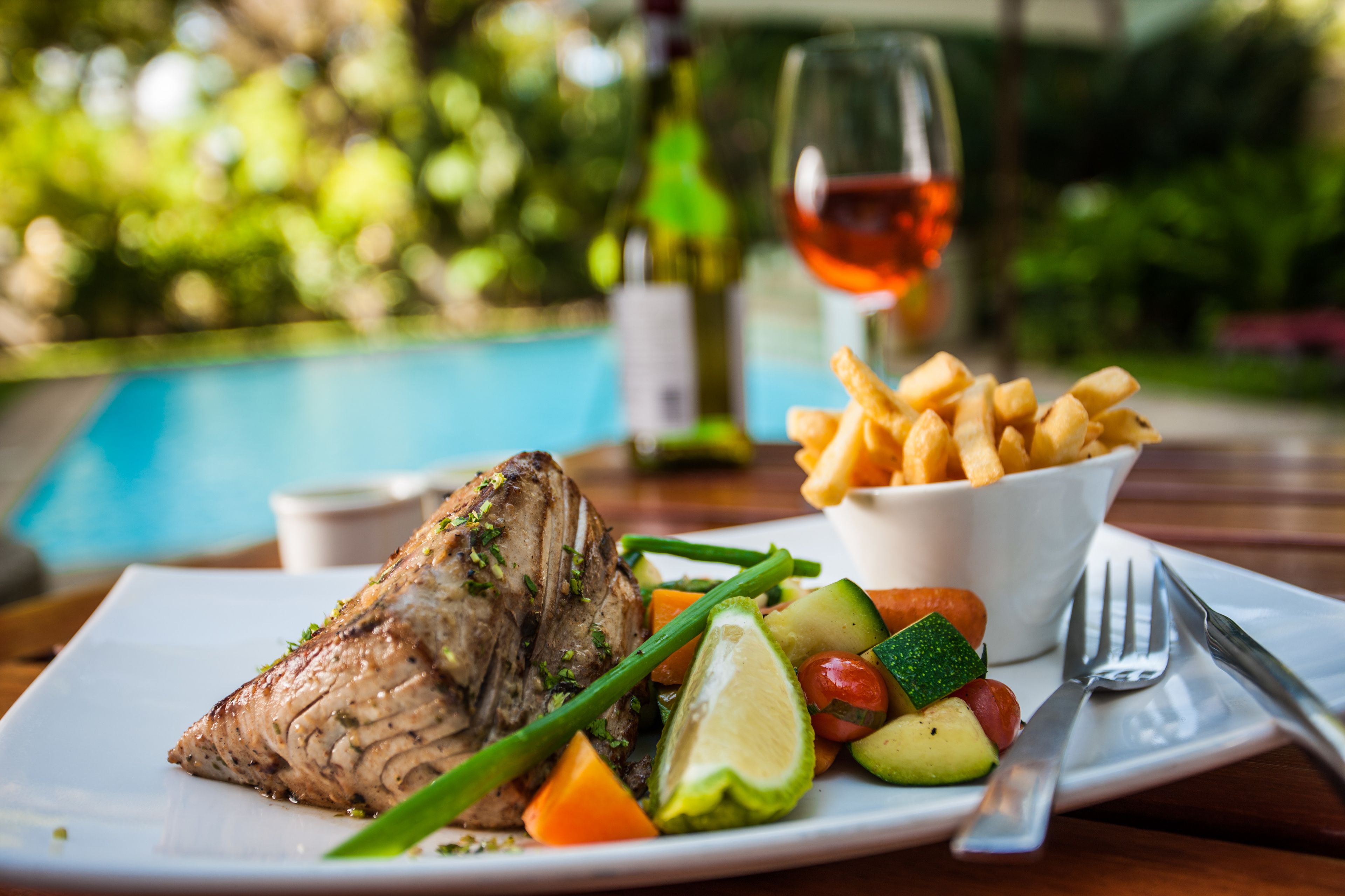 Un plato con carne a la parrilla, verduras y papas fritas, acompañado por una copa de vino tinto, dispuesto al aire libre junto a una piscina.