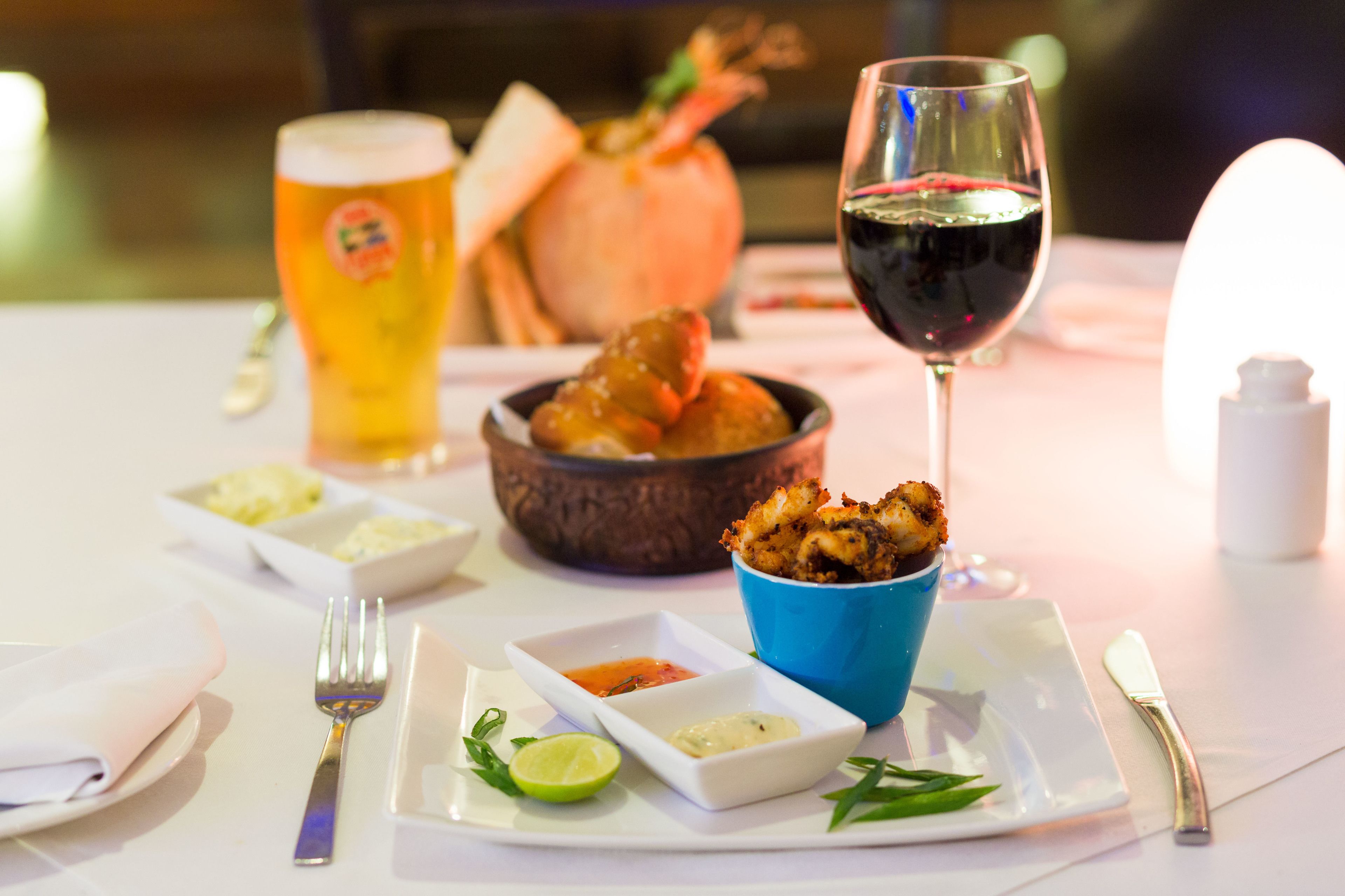 Una mesa de comedor con un plato de comida, un bol de camarones, una copa de vino tinto y una jarra de cerveza.