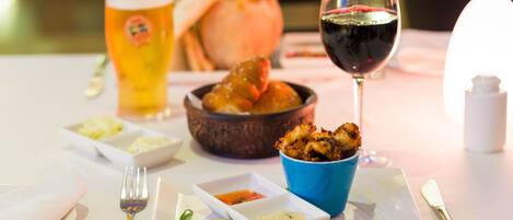 Una mesa de comedor con un plato de comida, un bol de camarones, una copa de vino tinto y una jarra de cerveza.