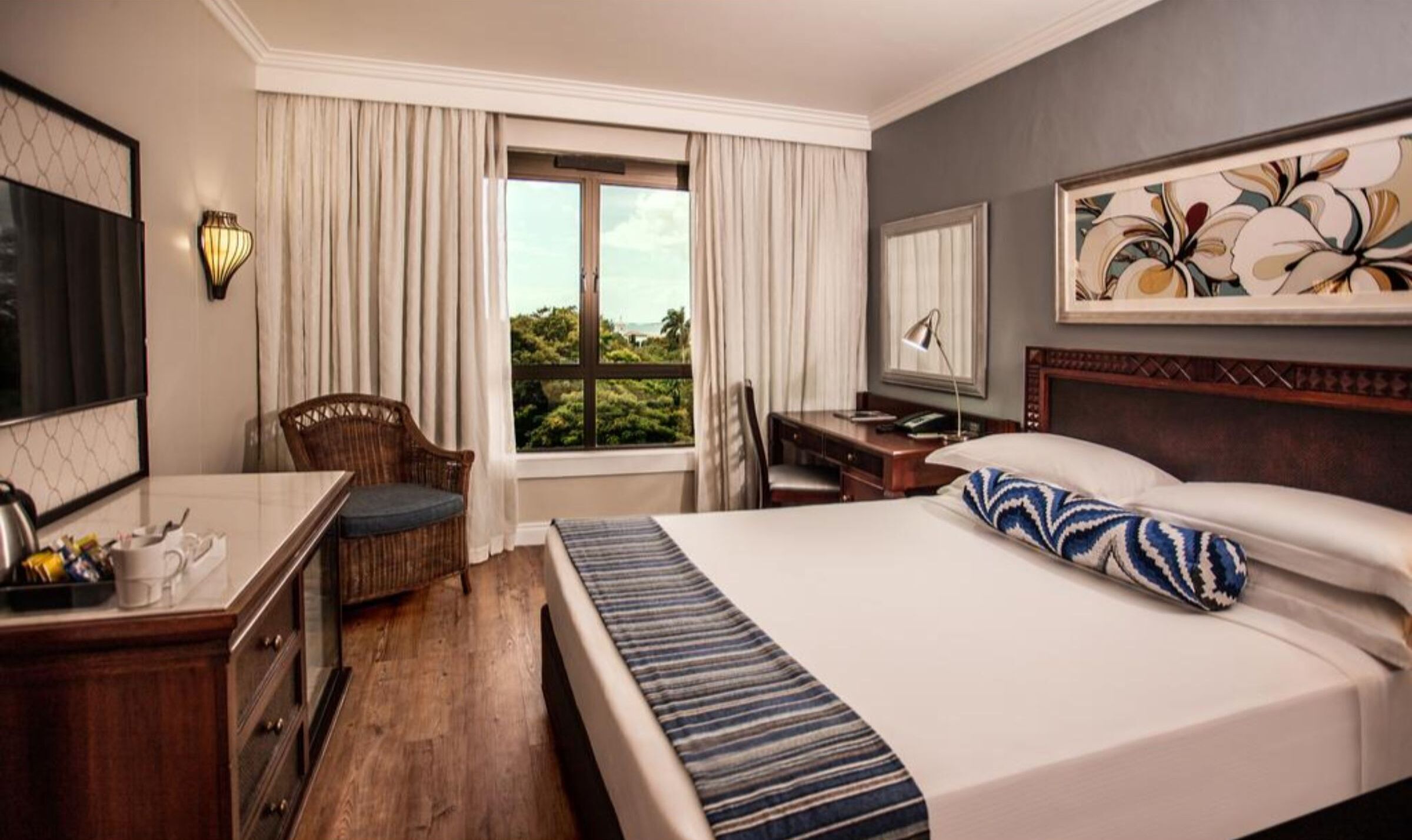 Une chambre d’hôtel avec un lit, un bureau, une chaise, une fenêtre avec des rideaux et un tableau au mur.
