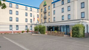 A hotel with a modern facade, multiple windows, and a covered outdoor seating area with benches. - Holiday Inn Express Dortmund by IHG (Dortmund)