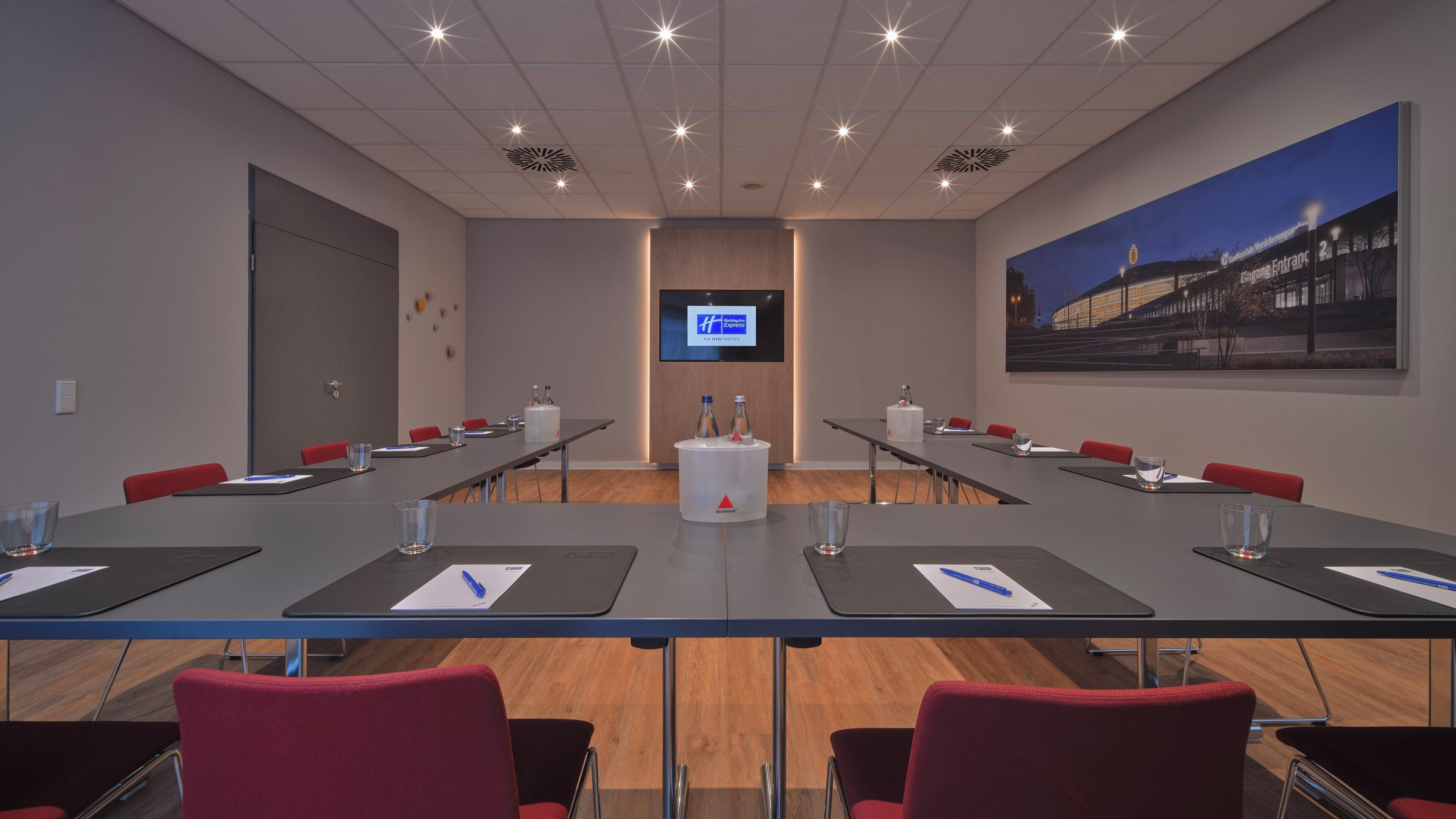 A conference room with a long table, red chairs, a large screen, and a painting on the wall.