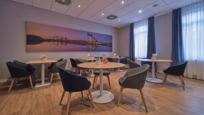 A modern dining area with round tables and chairs, a large cityscape painting on the wall, and a view of the outdoors through large windows. - Holiday Inn Express Dortmund by IHG (Dortmund)